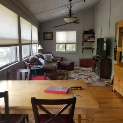 A cozy living room with a wooden dining table, chairs, couch, rugs, and a TV on a stand, lit by natural light through large windows.