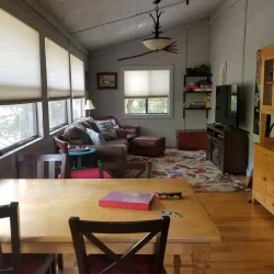 A cozy living room with a wooden dining table, chairs, couch, rugs, and a TV on a stand, lit by natural light through large windows.