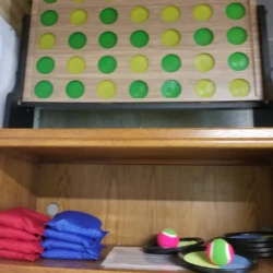 Wooden shelf with red and blue bean bags, discs, and two balls; a board with green and yellow circles leans above.