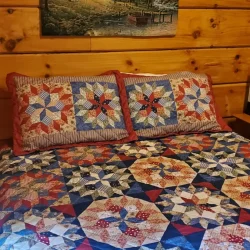 Quilt with intricate patterns on a wooden bed, below a rustic cabin painting on a wood-paneled wall.