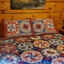 Quilt with intricate patterns on a wooden bed, below a rustic cabin painting on a wood-paneled wall.