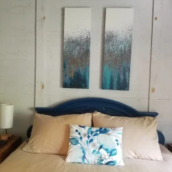 A neatly made bed with beige linens and a blue accent pillow, flanked by two lamps and two abstract paintings above the headboard.