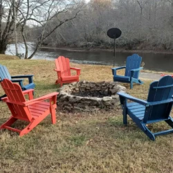 Six red and blue Adirondack chairs surround a stone fire pit beside a river, with bare trees in the background.