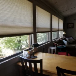 A cozy living room with large windows covered by blinds, a wooden table and chairs, a sofa, and a lamp.