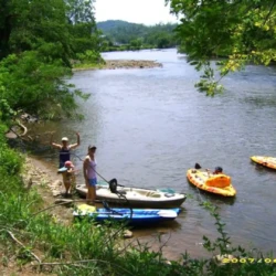 People at a riverbank with kayaks, some standing on the shore and others paddling in the water under a clear sky.
