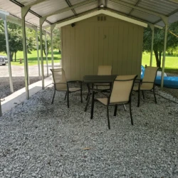 Outdoor seating area with a table and four chairs on gravel under a metal canopy. A shed and a blue kayak are in the background.