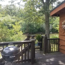 Wooden deck with a round barbecue grill, potted plants, a floral wreath on the wall, and a view of lush trees.