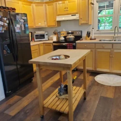 A kitchen with wooden cabinets, black appliances, a window, and a wheeled island with a bowl of fruit under a light fixture.