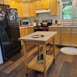 A kitchen with wooden cabinets, black appliances, a window, and a wheeled island with a bowl of fruit under a light fixture.