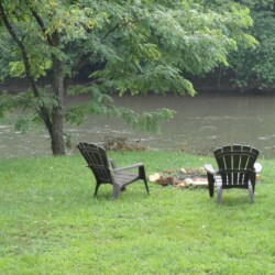 Two empty chairs face a river, surrounded by green grass and trees. A small fire pit is between the chairs.