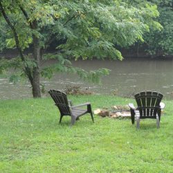 Two empty chairs face a river, surrounded by green grass and trees. A small fire pit is between the chairs.
