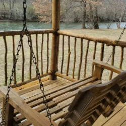 Wooden porch swing on a log cabin deck overlooking a grassy area and a river, surrounded by leafless trees.