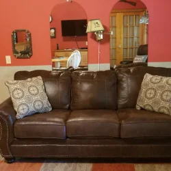 A brown leather couch with patterned pillows is set against a red and beige wall in a living room with wooden flooring.