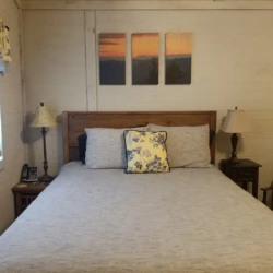 A cozy bedroom with a large bed, flanked by two lamps. A window is on the left, and three framed pictures hang above the bed.