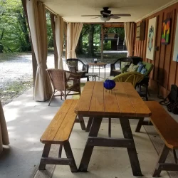 Covered patio with picnic table, wicker seating, ceiling fan, and artwork on the walls, surrounded by trees and gravel path.