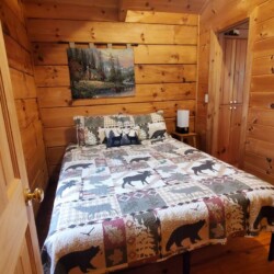 A cozy wooden cabin bedroom with a bear-themed quilt on the bed, a nature painting on the wall, and a small lamp on the nightstand.