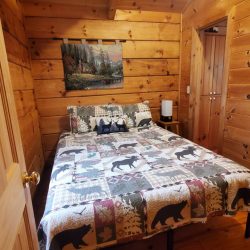 A cozy wooden cabin bedroom with a bear-themed quilt on the bed, a nature painting on the wall, and a small lamp on the nightstand.