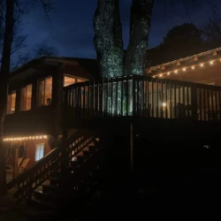 A wooden deck with string lights is attached to a house at night, surrounded by trees and a dark, cloudy sky.