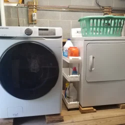A laundry room with a front-loading washing machine, a dryer on a wooden platform, and a basket and cleaning supplies on top.