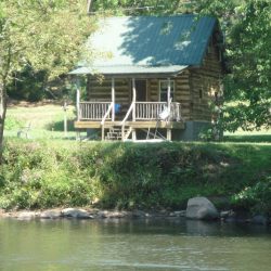 A rustic log cabin with a green roof is situated by a river, surrounded by trees and greenery.