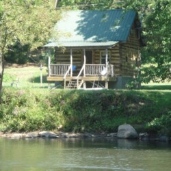 A rustic log cabin with a green roof is situated by a river, surrounded by trees and greenery.