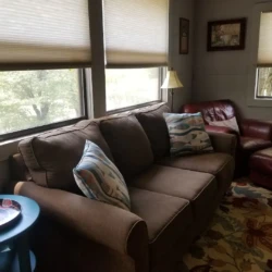 Cozy living room with a brown sofa, patterned pillows, a red armchair, footstool, and blue side table near large windows.