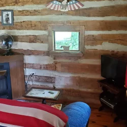 Cozy log cabin interior with a small window, television, electric fireplace, and a couch draped with an American flag-themed blanket.