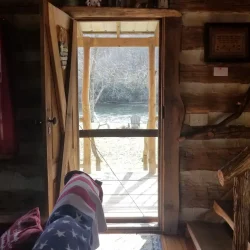 Interior of a log cabin with an open door leading to a sunlit porch, revealing a view of nature outside.