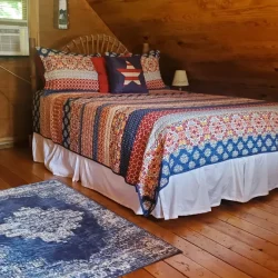 A cozy bedroom with a quilted bedspread, wooden walls, an air conditioner, a small lamp, and a blue patterned rug.