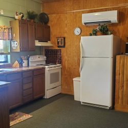 A cozy kitchen with wood paneling, a white fridge, stove, microwave, sink, cabinets, and a window with floral curtains.
