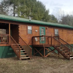 A small wooden cabin with a green metal roof and porch, located in a wooded area with another cabin visible in the background.