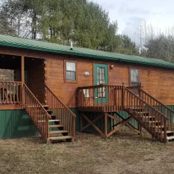 A small wooden cabin with a green metal roof and porch, located in a wooded area with another cabin visible in the background.