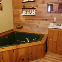 A rustic bathroom with a green corner bathtub, wooden paneling, a small sink, and shelves with nautical decorations and "DREAM" sign.