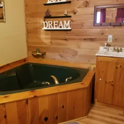 A rustic bathroom with a green corner bathtub, wooden paneling, a small sink, and shelves with nautical decorations and "DREAM" sign.