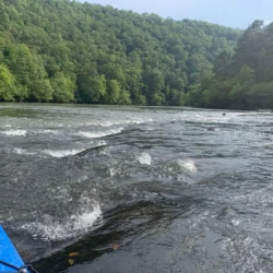A blue kayak on a river surrounded by lush, green forested hills under a clear sky.