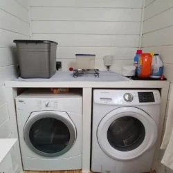 Laundry room with a washer and dryer beneath a counter, detergent bottles and a bin on top, in a white paneled space.
