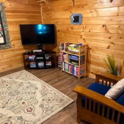 Cozy room with wood paneling, TV, bookshelf with games, patterned rug, armchair with a pillow, and a quilt on the wall.