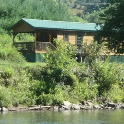 A small wooden cabin with a green roof is situated near a river, surrounded by trees and greenery.