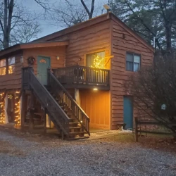 A wooden cabin with exterior stairs is adorned with string lights, surrounded by trees and a gravel driveway in a dim setting.