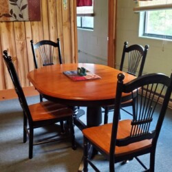 A wooden dining table with four chairs is set in a room with wood-paneled walls, a painting, and two windows with curtains.