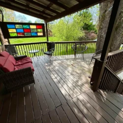 Deck with wooden railing, red cushioned sofa, chairs, and a colorful stained glass panel overlooking a grassy yard.