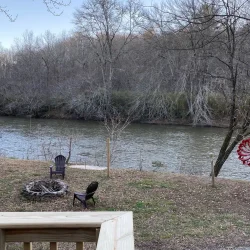Deck view of a river with bare trees, chairs around a fire pit, a satellite dish, and a decorative red windmill in the yard.