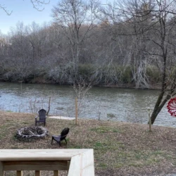 Deck view of a river with bare trees, chairs around a fire pit, a satellite dish, and a decorative red windmill in the yard.