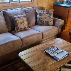 A cozy living room with a tan sofa, decorative pillows, a wooden coffee table, magazines, and a green cushioned chair.