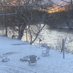 Three empty chairs surround a fire pit on a snow-covered riverside at dawn, with bare trees and a pastel sky in the background.
