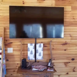 A flat-screen TV mounted on a wooden wall above a small table with a phone and modem. A quilt ladder is on the left.