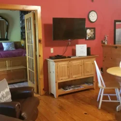 Cozy living area with red walls, wooden furniture, a wall-mounted TV, and a small dining table. Bedroom visible through an open door.