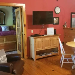 Cozy living area with red walls, wooden furniture, a wall-mounted TV, and a small dining table. Bedroom visible through an open door.