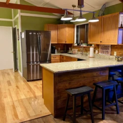 A kitchen with wooden cabinets, an island with granite countertop, black stools, stainless steel refrigerator, and hardwood floors.