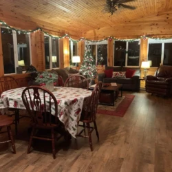 A cozy wooden interior features a decorated Christmas tree, dining table with festive cloth, and comfortable seating arrangements.
